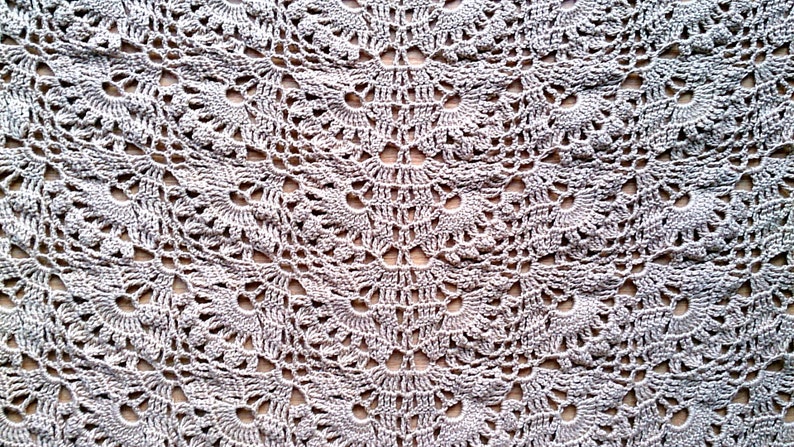 May include: Close-up of a crocheted textile with a repeating floral pattern. The handmade piece is in a neutral, light gray color, with a delicate, openwork design. The texture is soft and intricate, suitable for a shawl or blanket.