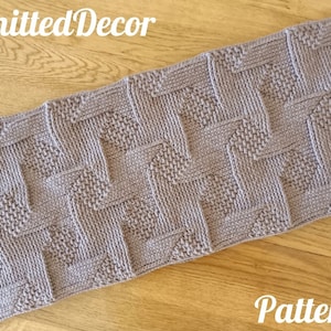 Può includere: Una sciarpa di lana grigia con un motivo geometrico. La sciarpa è distesa su una superficie di legno. Il testo "Knitted Decor" e "Pattern" è visibile nell'immagine.