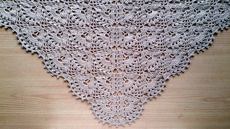 May include: A beige, handmade crochet shawl with a detailed, openwork pattern. The shawl is triangular in shape, with a scalloped edge. The intricate design features repeating floral-like motifs. The shawl is displayed on a light brown wooden surface.