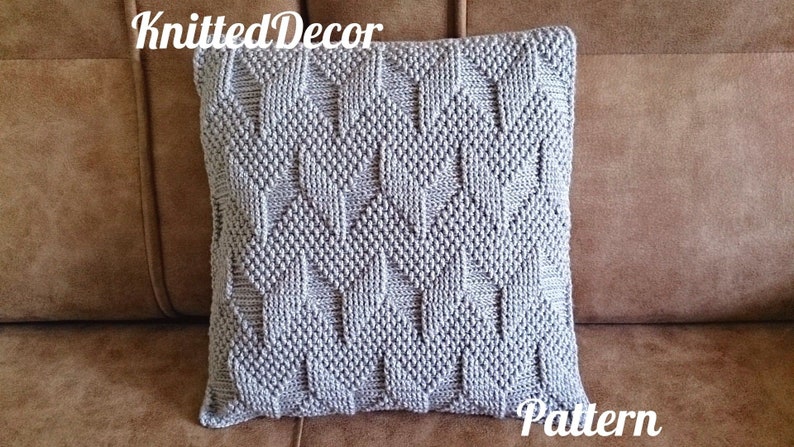 May include: A square, light grey knitted cushion with a textured geometric pattern. The cushion is on a brown surface. The text "Knitted Decor" and "Pattern" are in the image.