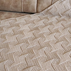 May include: A beige knitted blanket with a geometric pattern. The blanket is folded over a light brown couch. The pattern consists of repeating zig-zag shapes and textured squares, creating a visually interesting design. The blanket appears soft and comfortable.