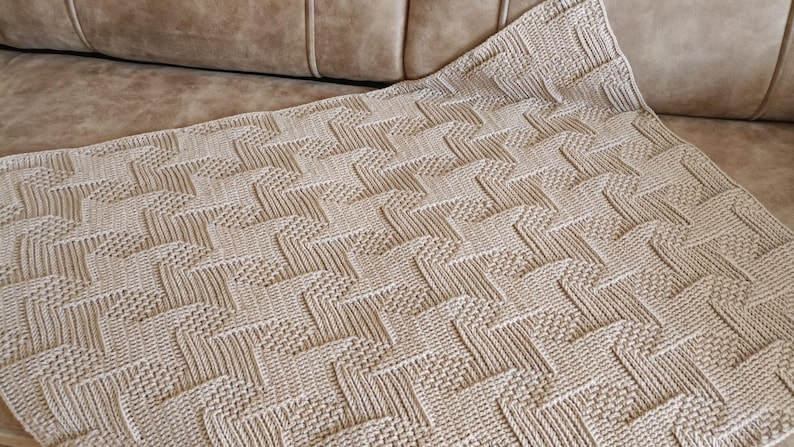 May include: A beige knitted blanket with a geometric pattern. The blanket is folded over a light brown couch. The pattern consists of repeating zig-zag shapes and textured squares, creating a visually interesting design. The blanket appears soft and comfortable.