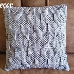 May include: A square, light grey knitted cushion with a geometric pattern. The cushion is placed on a brown surface. The text "KnittedDecor" is in the upper left corner and "Pattern" is in the lower right corner.