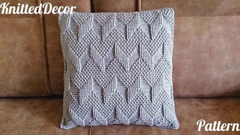 May include: A square, light grey knitted cushion with a geometric pattern. The cushion is placed on a brown surface. The text "KnittedDecor" is in the upper left corner and "Pattern" is in the lower right corner.