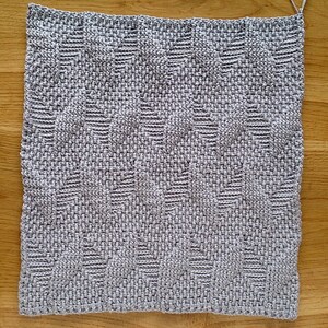 May include: A square, hand-woven textile in shades of grey, featuring a repeating geometric pattern. The texture appears soft and the weave is tight. The piece is likely a decorative item, such as a placemat or small rug.