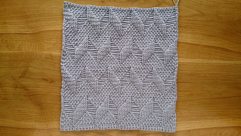 May include: A square, hand-woven textile in shades of grey, featuring a repeating geometric pattern. The texture appears soft and the weave is tight. The piece is likely a decorative item, such as a placemat or small rug.