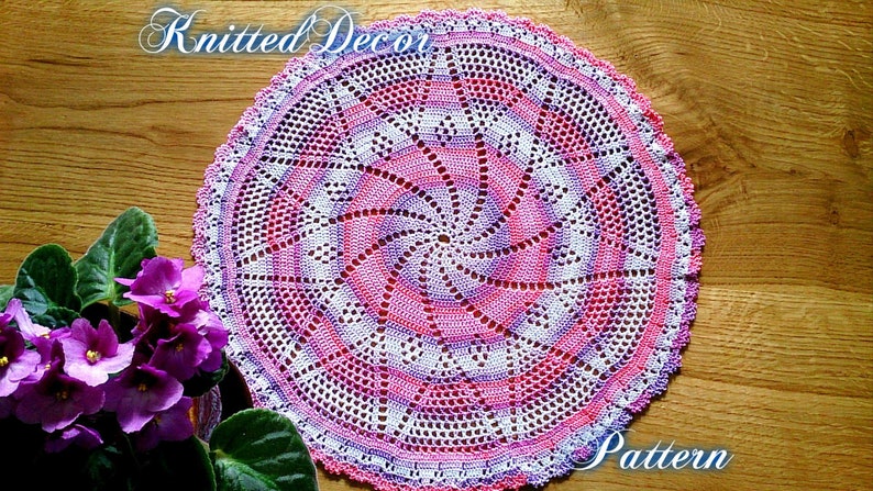 Puede incluir: Un tapete circular hecho a mano, con un dise&ntilde;o en espiral en tonos rosa, blanco y morado. El tapete est&aacute; tejido a ganchillo con un delicado patr&oacute;n calado y tiene un borde festoneado. Las palabras "Knitted Decor" y "Pattern" est&aacute;n escritas en la imagen.