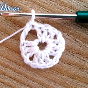Pu&ograve; includere: Primo piano di un cerchio bianco all'uncinetto con un uncinetto e filo bianco. L'uncinetto ha un manico verde. Il testo "Knitted Decor" &egrave; nell'angolo in alto a sinistra. Lo sfondo &egrave; una superficie di legno.