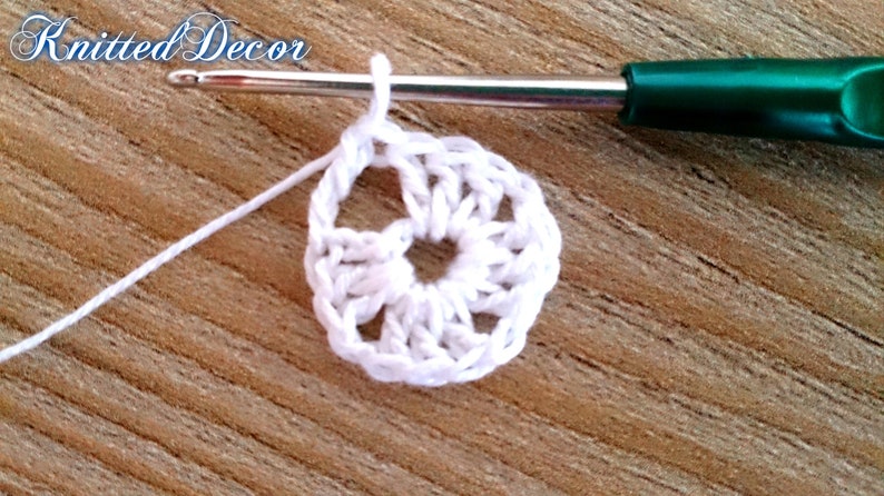 Pu&ograve; includere: Primo piano di un cerchio bianco all'uncinetto con un uncinetto e filo bianco. L'uncinetto ha un manico verde. Il testo "Knitted Decor" &egrave; nell'angolo in alto a sinistra. Lo sfondo &egrave; una superficie di legno.