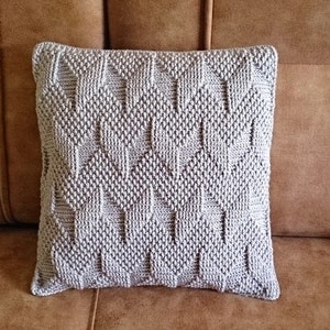 May include: A square, light grey decorative cushion with a textured, geometric pattern. The cushion is made of a soft material and is approximately 45 cm square. It is placed on a brown leather sofa.