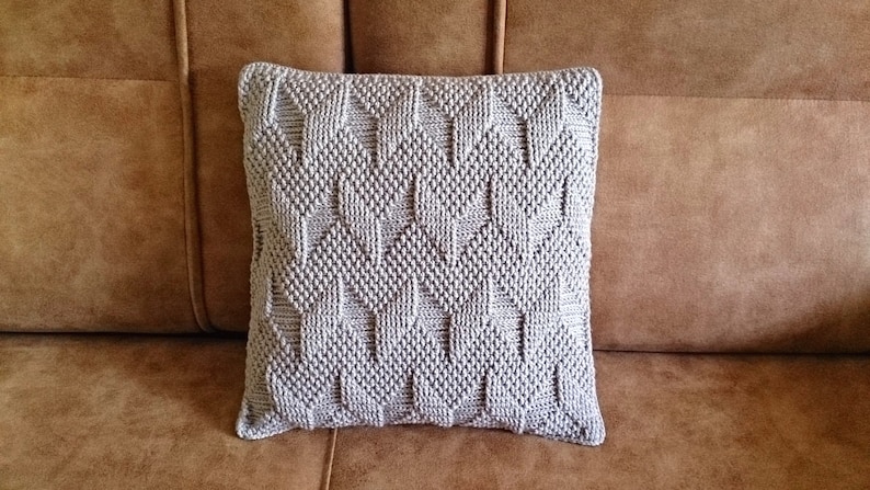 May include: A square, light grey decorative cushion with a textured, geometric pattern. The cushion is made of a soft material and is approximately 45 cm square. It is placed on a brown leather sofa.