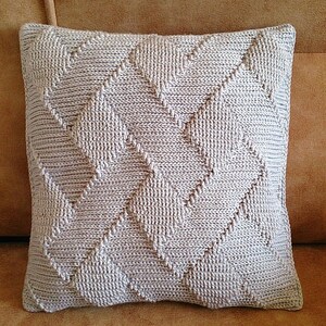 May include: A square throw cushion featuring a light beige knit cover with a prominent diagonal chevron or herringbone texture pattern. The fabric displays a consistent, repeating geometric stitch design across the entire front surface of the cushion.