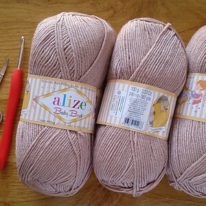 May include: Three balls of light brown yarn with labels, a red-handled crochet hook, and small scissors are arranged on a wooden surface. The yarn labels read "Alize Baby Best".