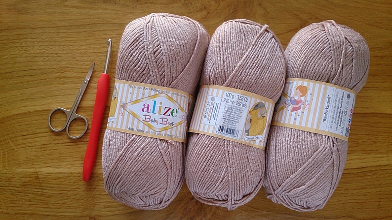May include: Three balls of light brown yarn with labels, a red-handled crochet hook, and small scissors are arranged on a wooden surface. The yarn labels read "Alize Baby Best".