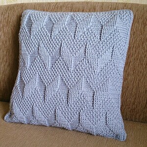 May include: A square, light grey knitted cushion with a textured geometric pattern. The cushion is placed on a brown sofa. The knitted design features a repeating diamond and rectangular pattern.