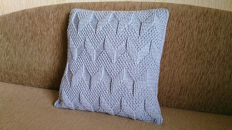 May include: A square, light grey knitted cushion with a textured geometric pattern. The cushion is placed on a brown sofa. The knitted design features a repeating diamond and rectangular pattern.