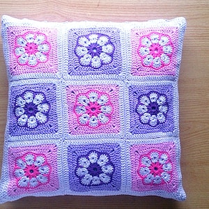 May include: A crocheted pillow with a floral pattern in shades of pink, purple, and white. The pillow is made of squares with a flower design in the center of each square.