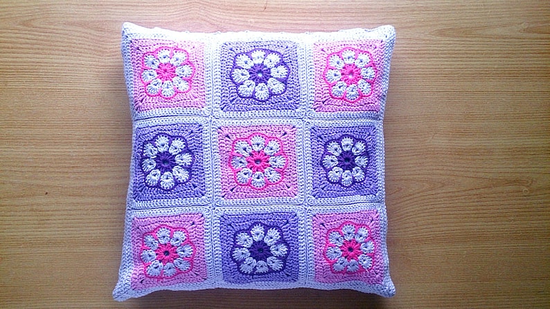 May include: A crocheted pillow with a floral pattern in shades of pink, purple, and white. The pillow is made of squares with a flower design in the center of each square.