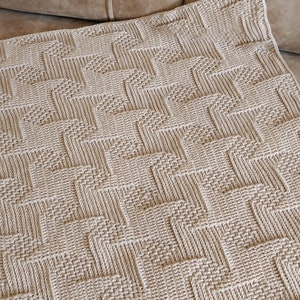 May include: A beige woven blanket with a geometric pattern. The blanket has a textured surface and is folded over a brown sofa. The pattern consists of repeating zig-zag shapes, creating a visually interesting design.