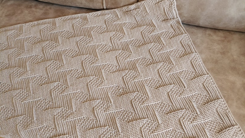 May include: A beige woven blanket with a geometric pattern. The blanket has a textured surface and is folded over a brown sofa. The pattern consists of repeating zig-zag shapes, creating a visually interesting design.