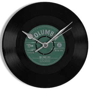 May include: Black vinyl record clock with a green label featuring the song "The Young Ones" by Cliff Richard and The Shadows. The label also includes the text "Columbia" and "45-DB 4781".