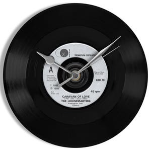 May include: A black vinyl record clock with silver clock hands. The clock face features the label of "Caravan of Love" by The Housemartins. The label includes the text "45 rpm" and the Warner Bros. Music Ltd. logo.