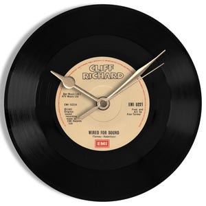 Op de afbeelding: Een zwarte vinylplaatklok met een crèmekleurig middenlabel met de tekst "CLIFF RICHARD" en "WIRED FOR SOUND". De wijzers zijn goudkleurig. De klok is rond en gemonteerd op een wit oppervlak.