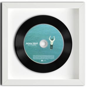 May include: A framed circular compact disc featuring the James Blunt album You're Beautiful. The disc has a black outer ring and a central image of a person jumping into blue water. The white frame is square and holds the disc centered.