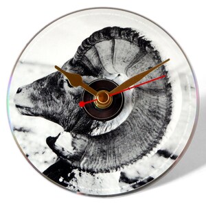 May include: A black and white image of a ram's head is printed on a white clock face. The clock has gold hands and a black center.
