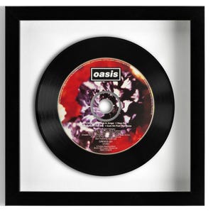 May include: A framed Oasis CD featuring a black disc with a red and purple floral graphic center label. The label displays the band name oasis in a white box, track titles including Don't Look Back in Anger, and production credits in white text.