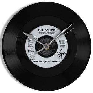 May include: A black vinyl record clock featuring the text "PHIL COLLINS" and "ANOTHER DAY IN PARADISE." The clock hands are silver, and the clock face includes the Virgin Records logo. The clock is a unique decorative item.