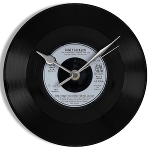 Op de afbeelding: Zwarte vinylplaat klok met een Janet Jackson label. De klokwijzers zijn zilverkleurig. Het label toont "WHAT HAVE YOU DONE FOR ME LATELY" en "45 RPM". Een originele wandklok voor muziekliefhebbers, ongeveer 30 cm in diameter.