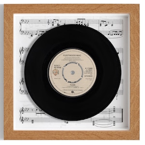 May include: A framed black vinyl record with a white label. The label has the text "Fleetwood Mac" and "Tusk" on it. The record is displayed on a sheet of music.