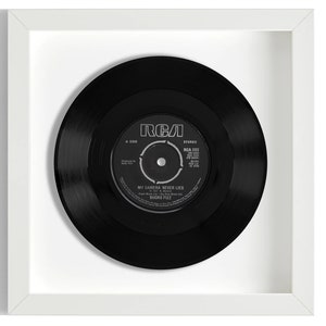 Peut inclure: Un disque vinyle noir dans un cadre blanc. L'étiquette du disque est RCA et le titre de la chanson est "My Camera Never Lies" de Buddy Fuzz.