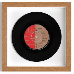 May include: A framed 7-inch vinyl record by The Wurzels. The record is black with a red label that says "I Am A Cider Drinker" and "The Wurzels". The record is framed in a light wood frame with a white mat.