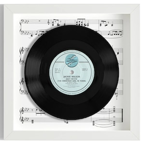 May include: A framed black vinyl record with a white label. The label has the text "Jackie Wilson - Sweet Petite (The Sweetest Girl In Town)" and "SKM 3". The record is displayed on a sheet of music.