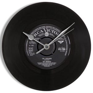 May include: Black vinyl record clock with silver hands. The clock face features the label of a 45 RPM record by The Monkees, titled "I'm a Believer." The clock has a classic, retro aesthetic.