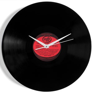 May include: A circular wall clock crafted from a black vinyl record featuring a central red label with the text mfp and Kenny Rogers. The clock has white hour and minute hands positioned over the record surface, which displays track listings.