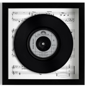 May include: A framed black vinyl record with the label "Roxy Music Dance Away" set against a backdrop of sheet music. The record is centered within a black frame, creating a striking contrast.