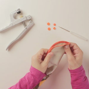 Op de afbeelding: Een persoon naait een stoffen tasje met een naaiwerktuig en oranje drukknopen. Het tasje is gemaakt van witte stof met een kleurrijk patroon. Er zijn oranje drukknopen en een naald op het witte oppervlak.