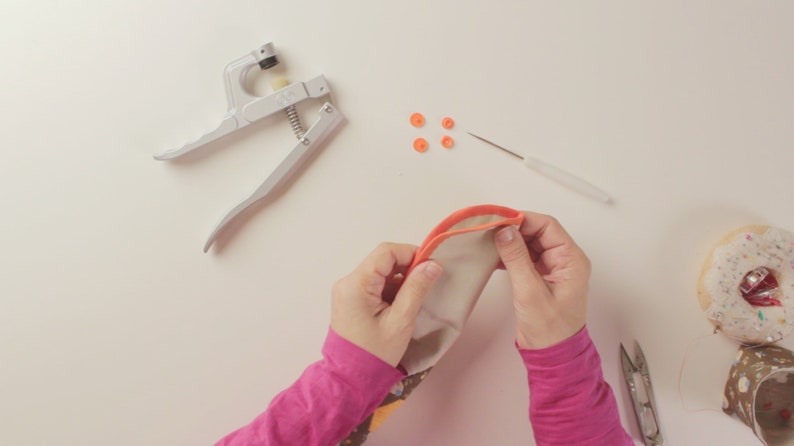 Op de afbeelding: Een persoon naait een stoffen tasje met een naaiwerktuig en oranje drukknopen. Het tasje is gemaakt van witte stof met een kleurrijk patroon. Er zijn oranje drukknopen en een naald op het witte oppervlak.