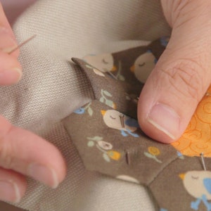 Op de afbeelding: Een close-up van een hand die een stoffen hexagon met een vogel- en bloemenpatroon vastpint op een groter stuk stof. De hexagon is omringd door andere stoffen hexagons met vergelijkbare patronen.