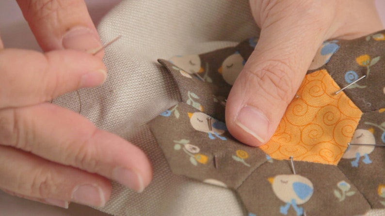 Op de afbeelding: Een close-up van een hand die een stoffen hexagon met een vogel- en bloemenpatroon vastpint op een groter stuk stof. De hexagon is omringd door andere stoffen hexagons met vergelijkbare patronen.