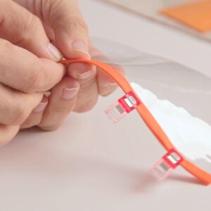 Op de afbeelding: Een doorzichtig plastic vel met een oranje rand wordt door een hand vastgehouden. Twee rode plastic clips zijn aan de oranje rand bevestigd.