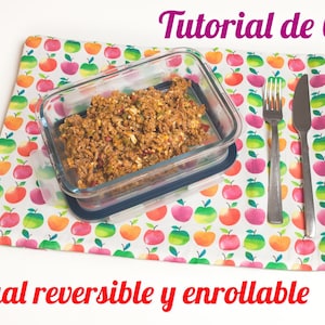 Puede incluir: Primer plano de un mantel de tela blanco con un patrón de manzanas rojas. Un recipiente de vidrio con comida en su interior está sobre el mantel. Un tenedor y un cuchillo están a la derecha del recipiente. El texto "Tutorial de Costura" está en la parte superior de la imagen. El texto "Individual reversible y enrollable" está en la parte inferior de la imagen. El texto "Mi Desenchufe" está en la esquina inferior derecha de la imagen.