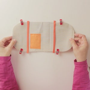 Op de afbeelding: Een witte stoffen tas met een klein oranje vakje wordt vastgehouden door twee handen. De tas wordt bij elkaar gehouden met rode clips.