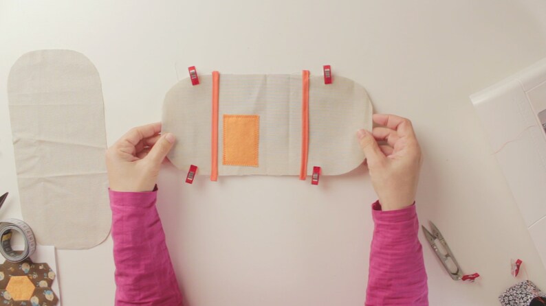 Op de afbeelding: Een witte stoffen tas met een klein oranje vakje wordt vastgehouden door twee handen. De tas wordt bij elkaar gehouden met rode clips.