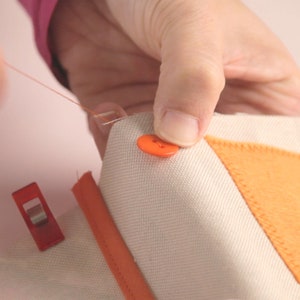 Op de afbeelding: Een close-up van een hand die een oranje knoop aan een stuk wit stof naait. De stof heeft een oranje viltplek die erop is genaaid en wordt op zijn plaats gehouden met rode clips.