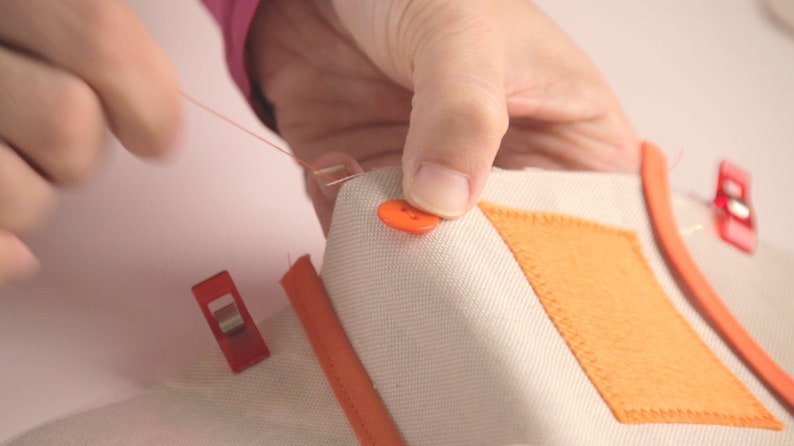 Op de afbeelding: Een close-up van een hand die een oranje knoop aan een stuk wit stof naait. De stof heeft een oranje viltplek die erop is genaaid en wordt op zijn plaats gehouden met rode clips.