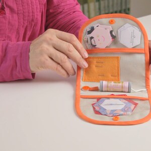 Op de afbeelding: Een open naaiset van beige stof met een oranje rand. De set bevat een spoel garen, naalden en stofresten.